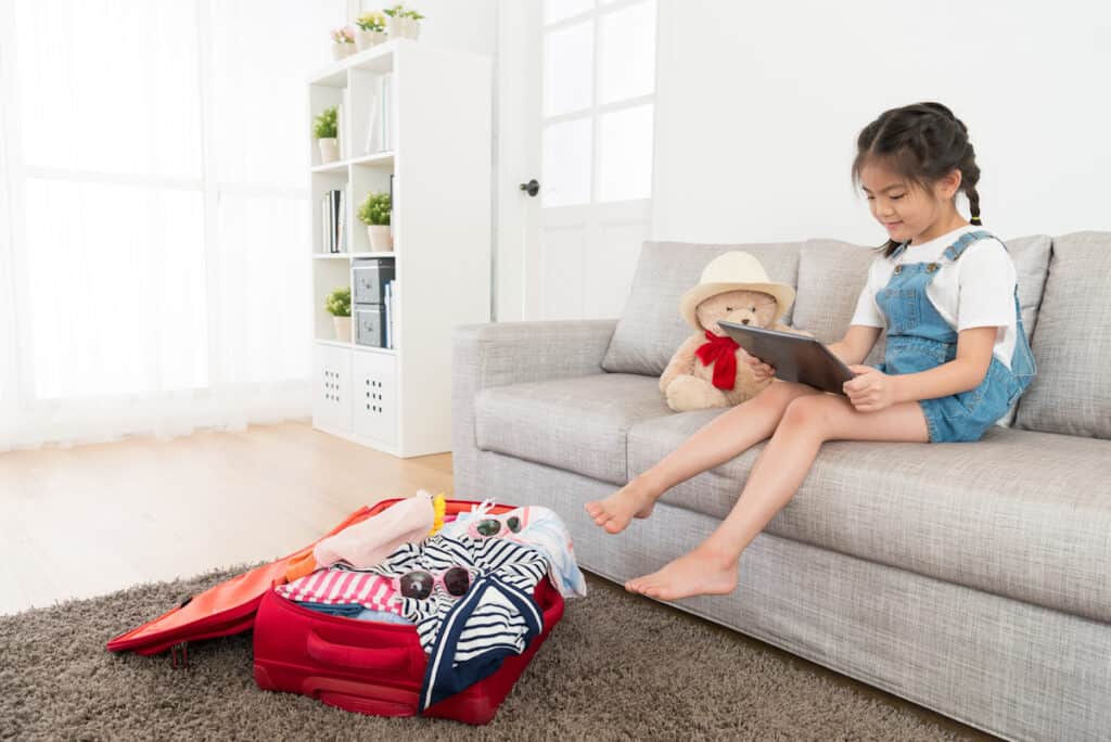 beautiful sweet kid girl sitting on living room sofa using mobile digital tablet pad searching travel information when she packing luggage suitcase at home.