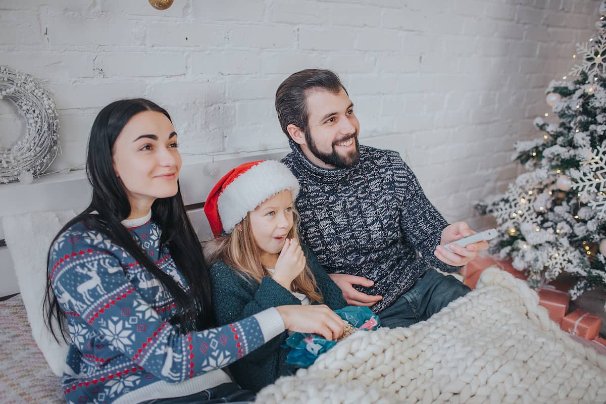 Merry Christmas and Happy New Year . Young family celebrating holiday at home. The Father is holding the remote from the TV. Dad, daughter and mother are watching television