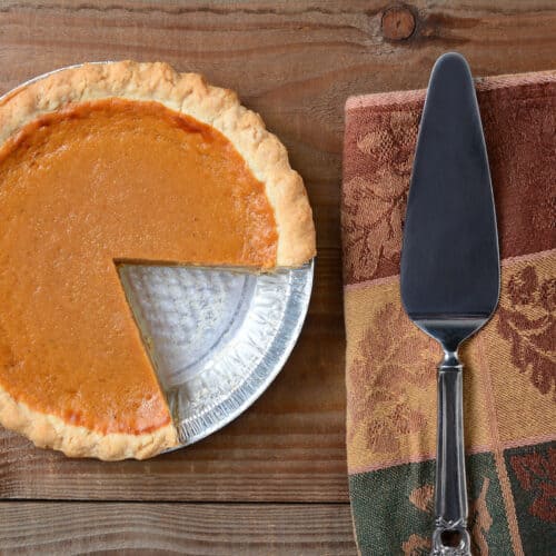 High angle shot of a pumpkin pie that has been cut into. Shot on a rustic wood table with a Autumn themed napkin and server by its side. Horizontal format.