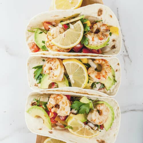 Seafood. Mexican food. Tortilla tacos with traditional homemade salsa salad, parsley, fresh lemon, avocado and grilled shrimp pawns. On a white marble background. Top view copy space